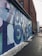A colorful mural with large letters spelling out an inspirational message adorns the side of a brick building. The mural uses shades of blue, green, and purple. Snow lines the edge of the sidewalk, and a person wearing a blue jacket and hat is walking along the path.