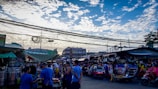 A bustling outdoor market scene with numerous people engaging in various activities. Vendors sell their goods under makeshift tents, while shoppers browse and purchase items. The sky is partly cloudy, with electric wires stretching across the scene, and several buildings are visible in the background.