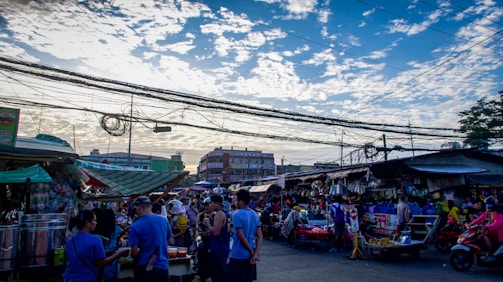 A bustling outdoor market scene with numerous people engaging in various activities. Vendors sell their goods under makeshift tents, while shoppers browse and purchase items. The sky is partly cloudy, with electric wires stretching across the scene, and several buildings are visible in the background.