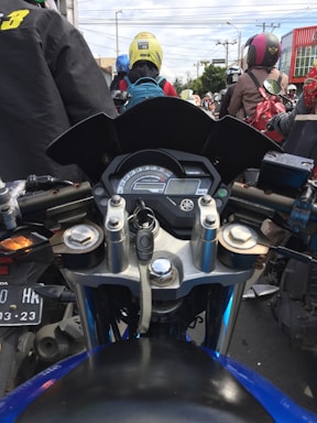 A view from the perspective of a motorcyclist in the midst of traffic. The motorcycle's handlebars and dashboard are in the foreground, with various vehicles and helmeted riders visible ahead. The riders are wearing different colored helmets and backpacks, and a building can be seen to the right.