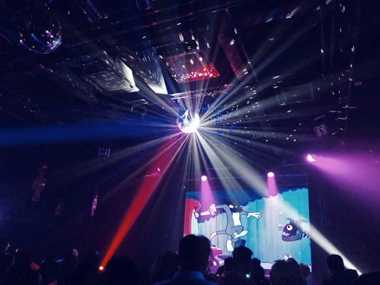A lively club scene with a disco ball casting light beams across the room, creating a dynamic pattern of light and shadow. In the background, a colorful animated projection is displayed, adding to the vibrant atmosphere. Silhouettes of people are visible, enjoying the music and ambiance.