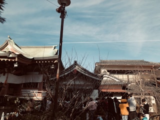 A traditional Asian architectural scene with wooden buildings and tiled roofs set against a clear blue sky. A group of people is gathered in the foreground, many wearing casual clothing. A leafless tree is present, and a vertical lamp post divides the scene.