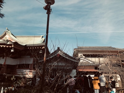 A traditional Asian architectural scene with wooden buildings and tiled roofs set against a clear blue sky. A group of people is gathered in the foreground, many wearing casual clothing. A leafless tree is present, and a vertical lamp post divides the scene.