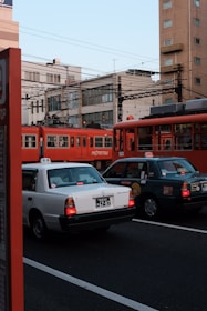 A city street scene with two taxis in the foreground, a white one and a dark-colored one, stopped at an intersection. In the background, two red tram cars are visible, with overhead electric lines running above. The scene is set against a backdrop of urban buildings and a clear sky.