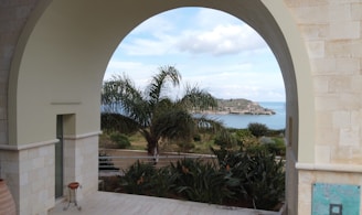 A large stone archway frames a scenic view of a coastal landscape with palm trees and an island in the distance. Potted plants decorate the foreground on a light-colored tiled floor, and there is a mixture of natural and architectural elements creating a serene environment.