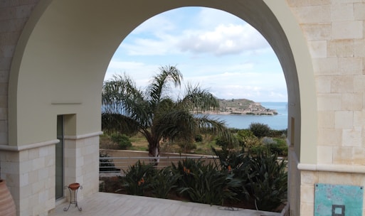A large stone archway frames a scenic view of a coastal landscape with palm trees and an island in the distance. Potted plants decorate the foreground on a light-colored tiled floor, and there is a mixture of natural and architectural elements creating a serene environment.