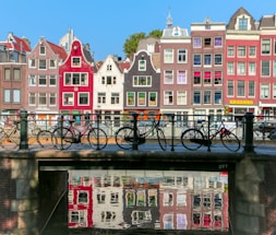 A row of colorful, traditional Dutch canal houses with stepped gables is reflected in the water of a canal. Several bicycles are parked along a metal railing on a bridge crossing the canal. The sky is clear and blue, suggesting a sunny day.