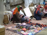 A group of people, likely vendors, are sitting on the ground beside a stone wall and selling various colorful goods. The vendors are adorned with bright, traditionally styled clothing and headwear decorated with tassels. The items for sale include intricately patterned textiles, small handcrafted boxes, and other assorted trinkets laid out on cloths.