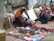 A group of people, likely vendors, are sitting on the ground beside a stone wall and selling various colorful goods. The vendors are adorned with bright, traditionally styled clothing and headwear decorated with tassels. The items for sale include intricately patterned textiles, small handcrafted boxes, and other assorted trinkets laid out on cloths.