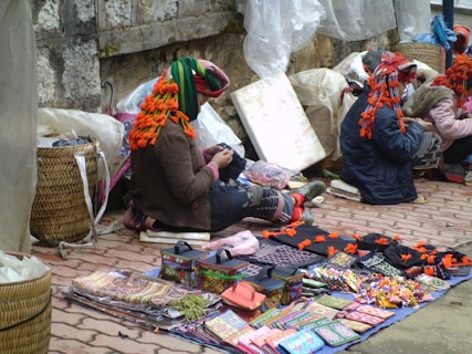 A group of people, likely vendors, are sitting on the ground beside a stone wall and selling various colorful goods. The vendors are adorned with bright, traditionally styled clothing and headwear decorated with tassels. The items for sale include intricately patterned textiles, small handcrafted boxes, and other assorted trinkets laid out on cloths.