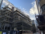 A construction site with extensive scaffolding surrounding a building under development. The scene captures the urban setting with modern architecture around. The sky has scattered clouds and the sunlight reflects off the building surfaces.