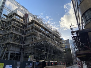 A construction site with extensive scaffolding surrounding a building under development. The scene captures the urban setting with modern architecture around. The sky has scattered clouds and the sunlight reflects off the building surfaces.