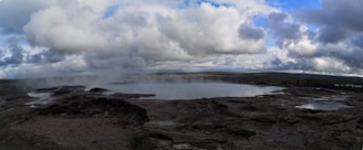 A geothermal landscape with a steaming pool surrounded by rocky terrain under a cloudy sky.