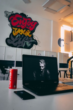 A room with a white interior features a colorful wall mural with the words 'Enjoy your life.' In the foreground, a Dell laptop displays a grayscale image of a serious-looking figure. Next to the laptop sits a red Coca-Cola can, and a smartphone lies on the table.