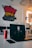 A room with a white interior features a colorful wall mural with the words 'Enjoy your life.' In the foreground, a Dell laptop displays a grayscale image of a serious-looking figure. Next to the laptop sits a red Coca-Cola can, and a smartphone lies on the table.