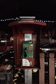 A vintage public payphone with a green and brown color scheme is positioned outdoors against a nighttime backdrop. The phone is encased in a shelter with the words 'Powered by PERNEC' displayed above. Nearby, there are muted streetlights and a faint silhouette of a busy road with passing vehicles, providing a sense of movement against the stillness of the phone booth.
