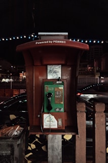 A vintage public payphone with a green and brown color scheme is positioned outdoors against a nighttime backdrop. The phone is encased in a shelter with the words 'Powered by PERNEC' displayed above. Nearby, there are muted streetlights and a faint silhouette of a busy road with passing vehicles, providing a sense of movement against the stillness of the phone booth.