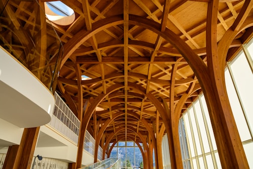 Intricate wooden arches and beams create an elaborate ceiling design in a modern architectural space. Floor-to-ceiling windows allow natural light to flood in, highlighting the warmth of the wood. The structure blends contemporary and natural elements, featuring geometric patterns and expansive views.