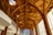 Intricate wooden arches and beams create an elaborate ceiling design in a modern architectural space. Floor-to-ceiling windows allow natural light to flood in, highlighting the warmth of the wood. The structure blends contemporary and natural elements, featuring geometric patterns and expansive views.