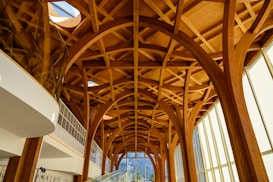 Intricate wooden arches and beams create an elaborate ceiling design in a modern architectural space. Floor-to-ceiling windows allow natural light to flood in, highlighting the warmth of the wood. The structure blends contemporary and natural elements, featuring geometric patterns and expansive views.