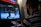 A person is playing a fighting video game on an arcade machine. The screen shows two characters engaged in a battle in a snowy setting. The person playing is focused, wearing glasses, and a striped shirt.