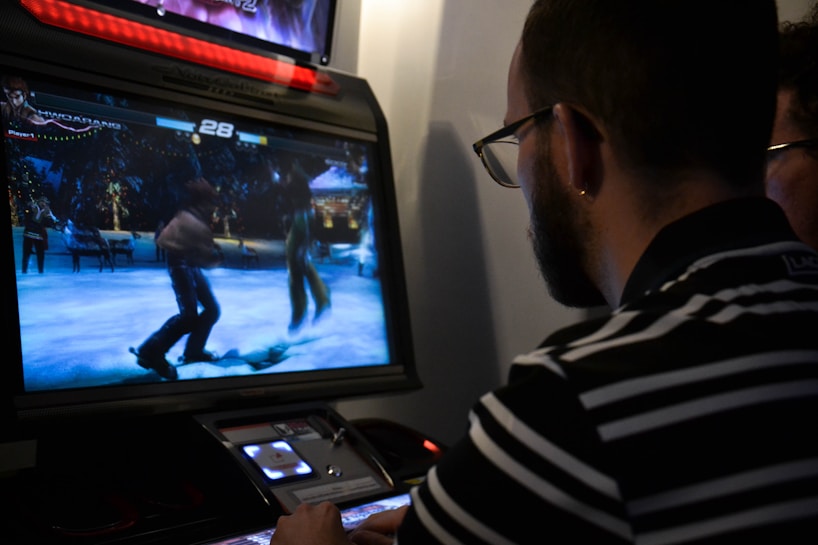 A person is playing a fighting video game on an arcade machine. The screen shows two characters engaged in a battle in a snowy setting. The person playing is focused, wearing glasses, and a striped shirt.