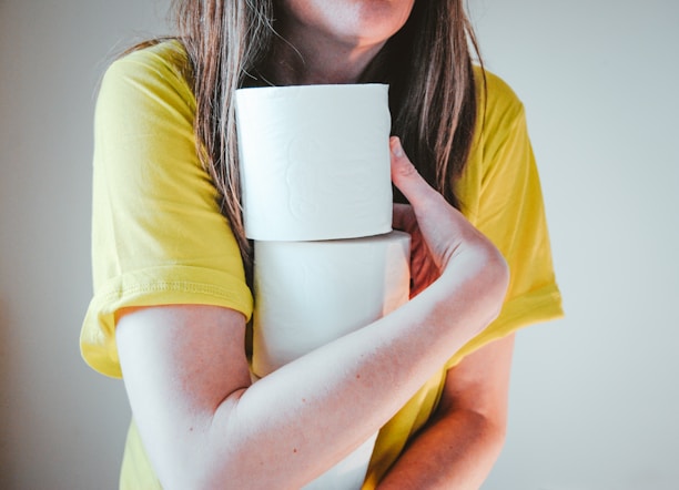 A person wearing a yellow short-sleeve shirt holds two rolls of toilet paper, cradling them in one arm. The focus is on the hands and the toilet paper, and the person's upper body is partially visible. The background is plain, adding emphasis to the subject.