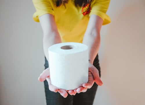 A person is wearing a yellow shirt and holding a roll of toilet paper with both hands. The focus is on the hands and the toilet paper.