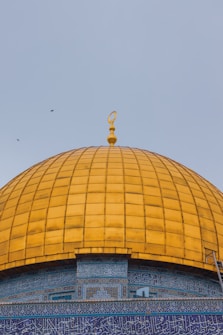 A large, domed structure with a shiny, golden surface is prominently displayed against a soft blue sky. The base of the dome features intricate blue and white tile work with Arabic script, adding to the architectural detail. A crescent finial adorns the top of the dome, enhancing its grandeur. Several birds are seen flying in the background.