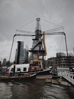 A large industrial crane structure stands prominently by the water, surrounded by modern buildings in the background. The crane has a numbered marking on it, and there is a boat named 'Volharding 1' docked nearby. Overcast skies add a somber tone to the scene.