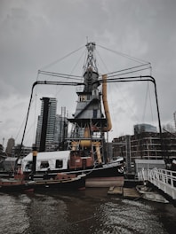 A large industrial crane structure stands prominently by the water, surrounded by modern buildings in the background. The crane has a numbered marking on it, and there is a boat named 'Volharding 1' docked nearby. Overcast skies add a somber tone to the scene.