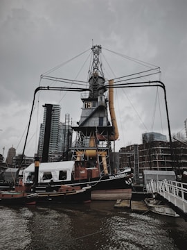 A large industrial crane structure stands prominently by the water, surrounded by modern buildings in the background. The crane has a numbered marking on it, and there is a boat named 'Volharding 1' docked nearby. Overcast skies add a somber tone to the scene.