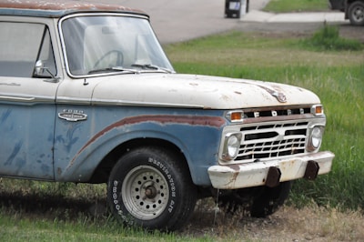 A vintage Ford truck with noticeable rust and wear, parked on grass. The vehicle features a mix of blue and white paint, with visible rust patches and aged metal parts. The truck's front grill and headlights are prominent, showcasing a classic design.