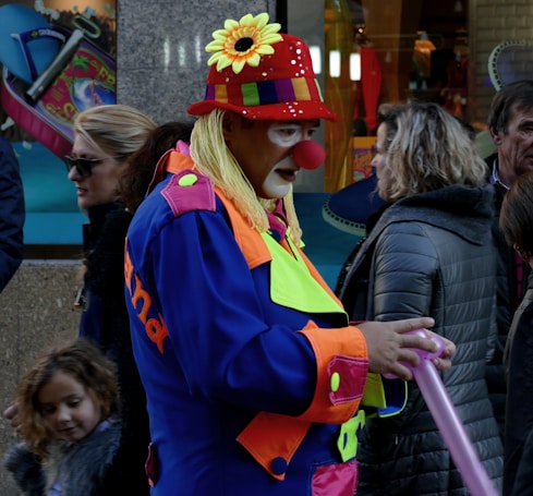 A clown in a colorful outfit interacts with people on a busy street. The clown is wearing a red hat with a sunflower, a big red nose, and a multicolored costume. Surrounding the clown are several people, including a child, all dressed in winter clothing. The background has shop windows displaying various items.