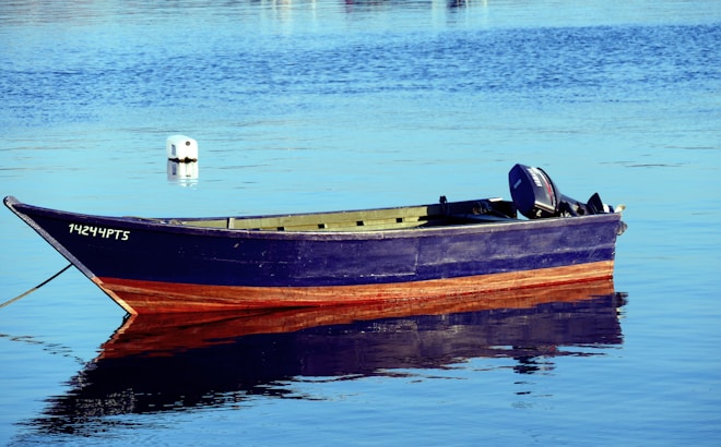 A small wooden boat with a dark blue and reddish-brown paint floats on calm water. It has an outboard motor attached to the back and a registration number on the side. The reflection of the boat is visible on the water's surface, and a white buoy is slightly visible in the background.