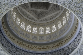 An intricately designed dome with detailed geometric and calligraphic patterns. The structure features an ornate repeating motif and a series of arched windows with decorative elements. Arabic script is prominently displayed, enhancing the architectural beauty and cultural richness.