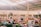 A bustling indoor marketplace featuring various stalls with colorful goods and fresh produce. People are interacting and shopping, with some seated and others walking around. The spacious area is lit by natural light filtering through a high, exposed ceiling.