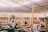 A bustling indoor marketplace featuring various stalls with colorful goods and fresh produce. People are interacting and shopping, with some seated and others walking around. The spacious area is lit by natural light filtering through a high, exposed ceiling.