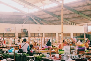 A bustling indoor marketplace featuring various stalls with colorful goods and fresh produce. People are interacting and shopping, with some seated and others walking around. The spacious area is lit by natural light filtering through a high, exposed ceiling.