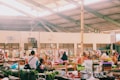 A bustling indoor marketplace featuring various stalls with colorful goods and fresh produce. People are interacting and shopping, with some seated and others walking around. The spacious area is lit by natural light filtering through a high, exposed ceiling.