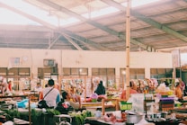 A bustling indoor marketplace featuring various stalls with colorful goods and fresh produce. People are interacting and shopping, with some seated and others walking around. The spacious area is lit by natural light filtering through a high, exposed ceiling.