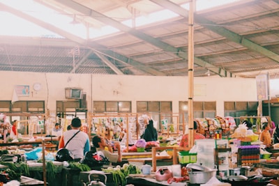 A bustling indoor marketplace featuring various stalls with colorful goods and fresh produce. People are interacting and shopping, with some seated and others walking around. The spacious area is lit by natural light filtering through a high, exposed ceiling.
