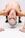 A muscular person is performing a physical pose, balancing with their hands on the ground while upside down. Their hair is styled with gray and white coloring, and a striking red line descends down their forehead and nose. They are shirtless, emphasizing their physique, and wearing black pants and black footwear. There is a tattoo visible on one of their forearms.