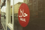 A red circular sign on a glass window displays the word 'HALAL' in white letters with Arabic script above it. The background shows a blurred street view with buildings.