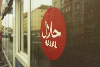 A red circular sign on a glass window displays the word 'HALAL' in white letters with Arabic script above it. The background shows a blurred street view with buildings.