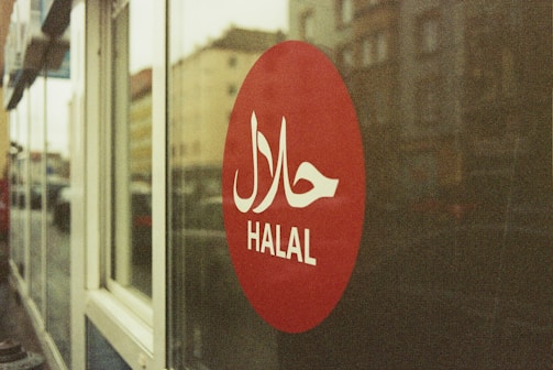 A red circular sign on a glass window displays the word 'HALAL' in white letters with Arabic script above it. The background shows a blurred street view with buildings.