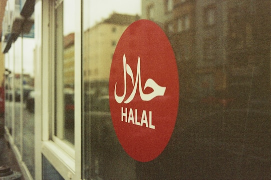 A red circular sign on a glass window displays the word 'HALAL' in white letters with Arabic script above it. The background shows a blurred street view with buildings.