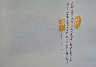 A piece of paper or parchment with vertical lines of Chinese calligraphy in dark ink. To the right of the script are two orange stamps or seals with traditional designs. The background is a muted off-white, and the calligraphy is meticulously arranged.