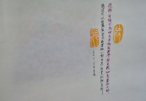 A piece of paper or parchment with vertical lines of Chinese calligraphy in dark ink. To the right of the script are two orange stamps or seals with traditional designs. The background is a muted off-white, and the calligraphy is meticulously arranged.