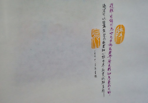 A piece of paper or parchment with vertical lines of Chinese calligraphy in dark ink. To the right of the script are two orange stamps or seals with traditional designs. The background is a muted off-white, and the calligraphy is meticulously arranged.